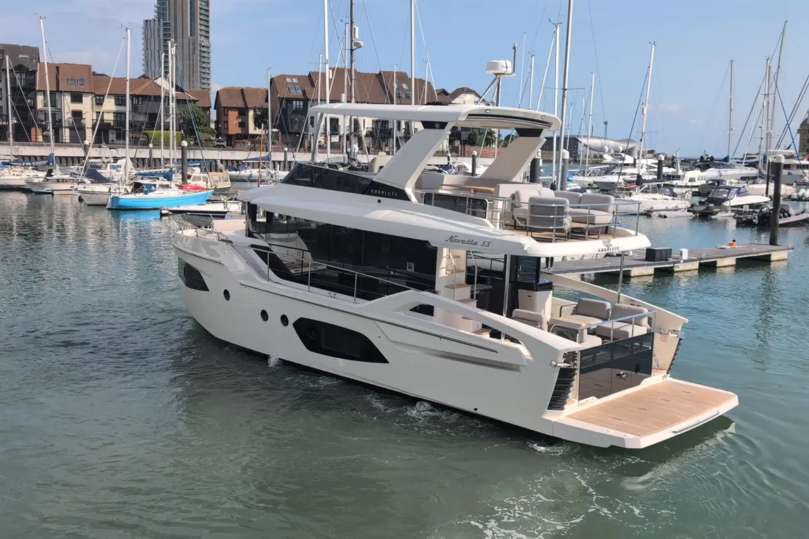 A sleek yacht named "Absolute Navetta 55" navigates a marina surrounded by smaller boats and modern buildings.